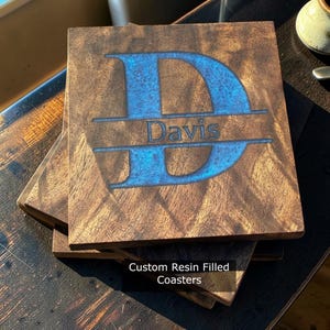 May include: Wooden square coasters with a blue resin-filled initial "D" and the name "Davis". The coasters are stacked, showcasing the wood grain and the custom design. The text "Custom Resin Filled Coasters" is at the bottom.