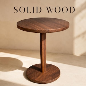 May include: A round side table crafted from dark brown solid wood, featuring a circular tabletop, a single central vertical support column, and a circular base. The text SOLID WOOD is displayed in dark brown capital letters above the table.