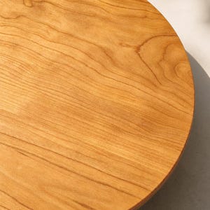 May include: A circular wooden board with a smooth, polished surface. The wood features a warm, light brown color with visible natural grain patterns running in parallel lines across the flat, round surface of the board.