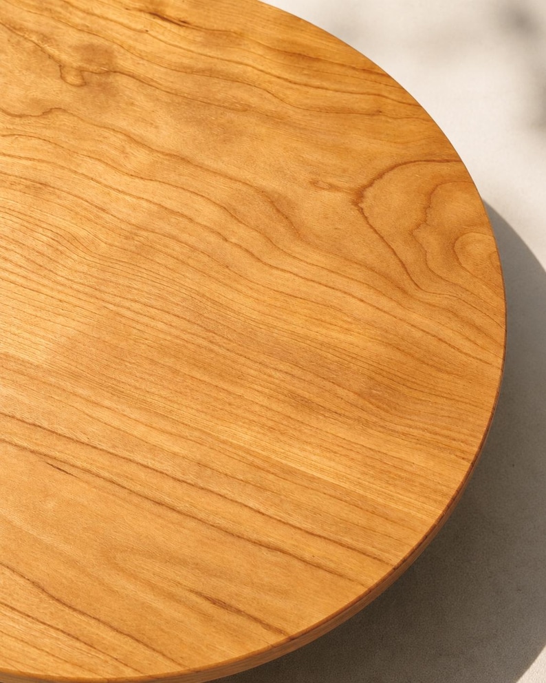 May include: A circular wooden board with a smooth, polished surface. The wood features a warm, light brown color with visible natural grain patterns running in parallel lines across the flat, round surface of the board.