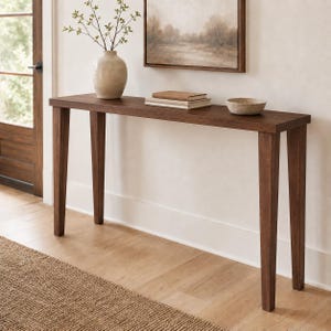 May include: A rectangular dark brown wooden console table with four tapered legs. On the surface sits a light-colored ceramic vase containing green branches, a stack of two beige books, and a small, shallow, round beige ceramic bowl.