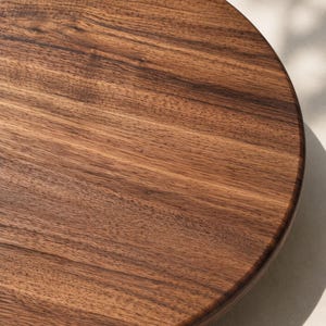 May include: A circular wooden board featuring a rich, dark brown grain pattern with visible natural wood textures. The surface is smooth and polished, showcasing the organic lines and deep color variations characteristic of natural walnut wood.