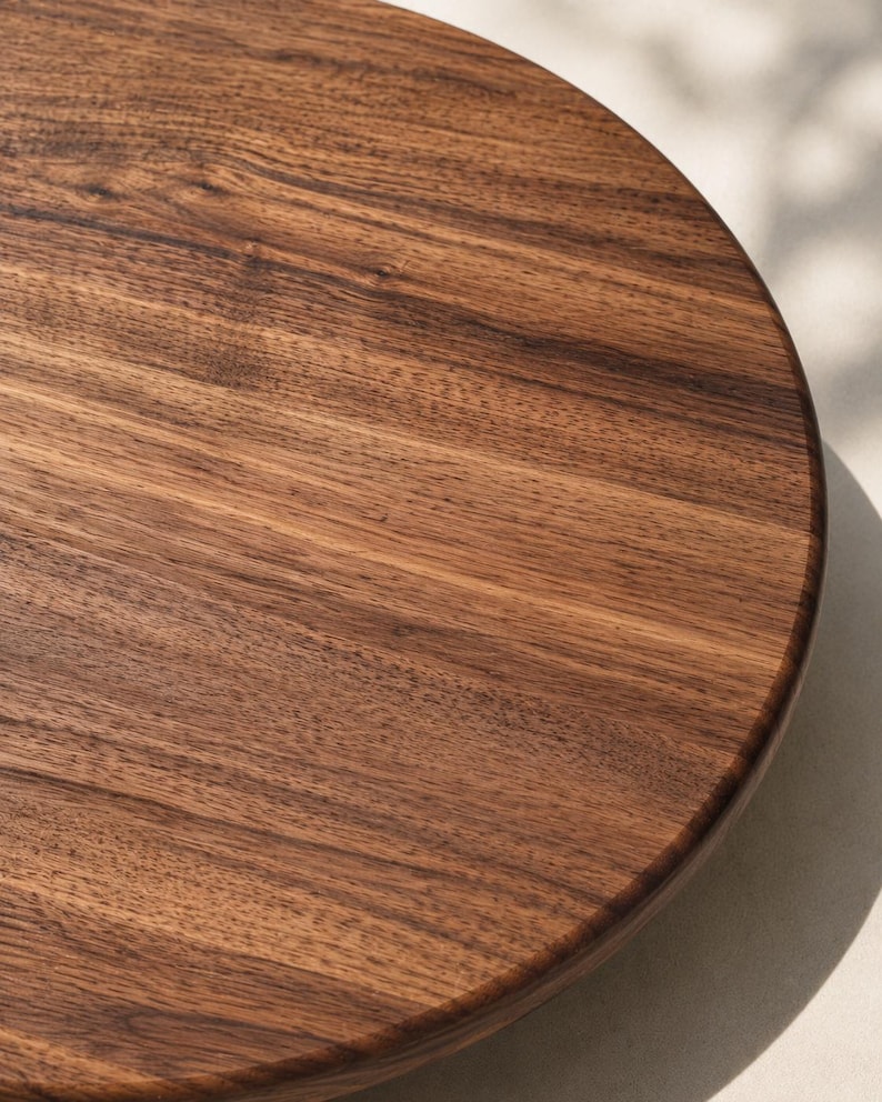 May include: A circular wooden board featuring a rich, dark brown grain pattern with visible natural wood textures. The surface is smooth and polished, showcasing the organic lines and deep color variations characteristic of natural walnut wood.
