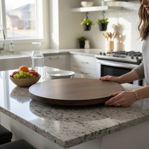 K&ouml;nnte beinhalten: Ein gro&szlig;es, rundes Serviertablett aus Holz mit dunkelbraunem Finish. Das Tablett wird auf einer Granit-Arbeitsplatte in einer K&uuml;che gehalten. Eine Sch&uuml;ssel mit Obst und ein Stapel Teller sind ebenfalls zu sehen.