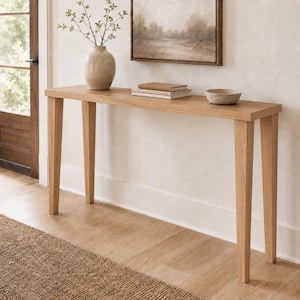 Può includere: Un tavolo console in rovere chiaro con quattro gambe affusolate. Sulla superficie rettangolare sono appoggiati un vaso in ceramica beige con rami verdi, una pila di due libri beige e una piccola ciotola beige, rotonda e poco profonda.