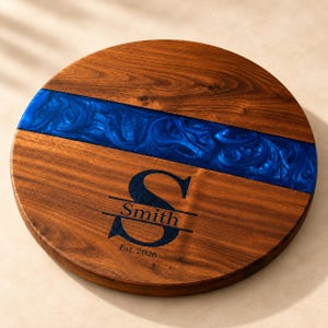 May include: A round wooden serving board featuring a dark brown wood grain and a horizontal band of swirling metallic blue resin. The board is engraved with a large letter S, the name Smith, and the text Est. 2026 centered on the wood surface.
