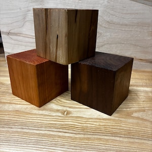 May include: Three wooden cubes in varying shades of brown, arranged in a stacked configuration. The wood grains are visible, with colors ranging from light tan to dark brown. The cubes are displayed on a light-colored wooden surface.
