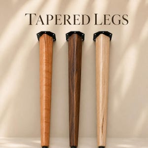Puede incluir: Tres patas de mueble cónicas hechas de madera en tonos variados de marrón claro, medio y oscuro. Cada pata cuenta con una placa de montaje de metal negro en la parte superior con orificios pretaladrados. El texto TAPERED LEGS aparece arriba.
