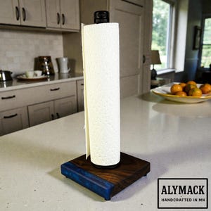 May include: A wooden paper towel holder with a dark wood base and a blue resin accent. The holder has a black top and holds a roll of white paper towels. The text "ALYMACK HANDCRAFTED IN MN" is in a white rectangle.