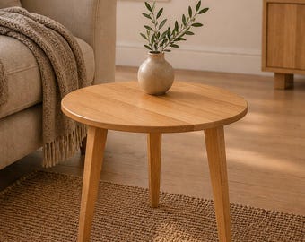 May include: A round wooden side table with three tapered legs, featuring a light oak finish and a smooth circular top. A small, textured beige ceramic vase containing a green olive branch sits centered on the surface of the table.