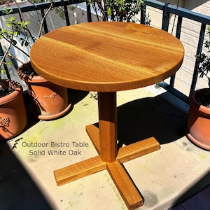 May include: A round outdoor bistro table made of solid white oak. The table has a natural wood finish and a pedestal base with four supporting legs. The text "Outdoor Bistro Table Solid White Oak" is visible.