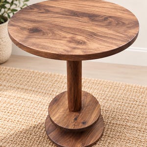 May include: A round wooden side table featuring a smooth, circular tabletop supported by a central cylindrical column. The base consists of two stacked circular wooden discs, all finished in a rich, dark brown wood grain pattern.