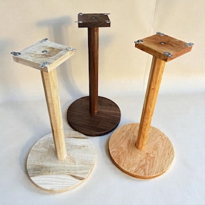 May include: Three wooden display stands with square tops and round bases. The stands are made of different types of wood, including light, medium, and dark tones. Each stand has metal corner brackets.