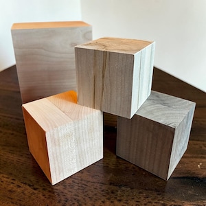 Peut inclure: Quatre blocs de bois de tailles et de formes variées. Le plus grand est un prisme rectangulaire, tandis que les autres sont des cubes. Le bois est de couleur claire naturelle avec des motifs de grain visibles. Les blocs sont disposés sur une surface en bois foncé.