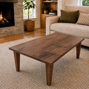 K&ouml;nnte beinhalten: Ein rechteckiger Couchtisch aus dunkelbraunem Holz mit einer glatten, flachen Oberfl&auml;che und vier stabilen, leicht konisch zulaufenden Beinen. Der Tisch weist eine sichtbare nat&uuml;rliche Holzmaserung auf der Oberseite auf.