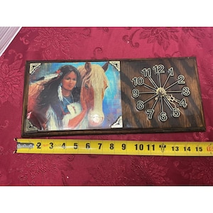 May include: A rectangular wooden clock featuring a Native American woman with a horse. The clock face has black numbers and gold hands. The clock is approximately 14 inches long.