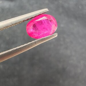 Peut inclure: Une pierre précieuse rose vif, de forme ovale, est maintenue par une pince argentée sur un fond gris foncé. Les facettes du joyau captent la lumière, révélant des textures internes et un éclat brillant.