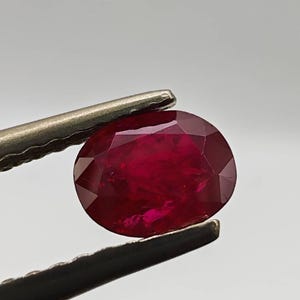 May include: A faceted, oval-shaped ruby gemstone held by tweezers. The ruby is a deep, saturated red color, with visible internal inclusions. The tweezers are silver-colored and hold the gemstone against a neutral background.