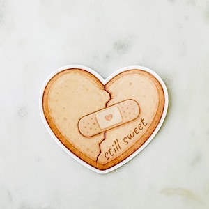 May include: A heart-shaped cookie sticker with a jagged crack down the center, held together by a tan adhesive bandage featuring a small heart icon. The text "still sweet" is printed in brown cursive script on the lower right side of the cookie.