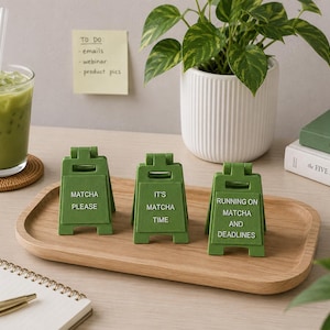 May include: Three small, olive green, A-frame sign displays made of plastic. Each sign features white text: the first reads MATCHA PLEASE, the second reads IT'S MATCHA TIME, and the third reads RUNNING ON MATCHA AND DEADLINES. The signs are standing upright.
