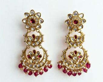 May include: A pair of gold-toned earrings with a floral design. Each earring features a flower-shaped top with a red center stone, followed by a crescent-shaped element with clear stones and red accents. Small red beads dangle from the bottom.