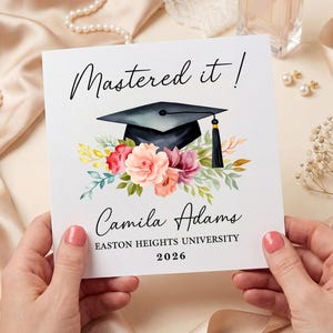 May include: A square graduation card held by hands, featuring the text "Mastered it!" above a mortarboard and floral arrangement. The card also includes the name "Camila Adams", university name, and the year 2026.