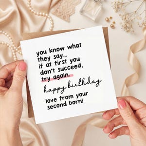 May include: A white birthday card with black text that reads "you know what they say... if at first you don't succeed, try again. happy birthday love from your second born!" The card is held by a person.