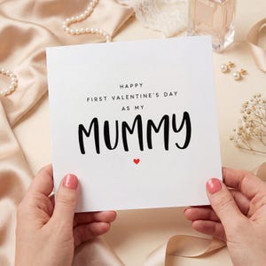 May include: A white card with the text "HAPPY FIRST VALENTINE'S DAY AS MY MUMMY" in black script, with a small red heart. The card is held by hands with pink nail polish.