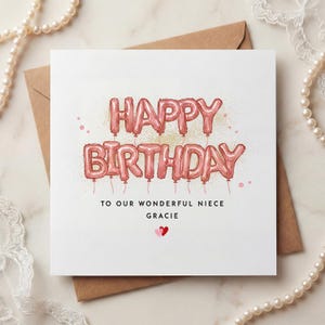 May include: A birthday card with pink balloon letters spelling "HAPPY BIRTHDAY". The text "TO OUR WONDERFUL NIECE GRACIE" is below, with two hearts. The card sits on a brown envelope, with pearls and lace in the background.