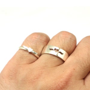 May include: Two silver rings with a heart design. The ring on the left has a small heart with wings, while the ring on the right has a larger heart with wings on either side.