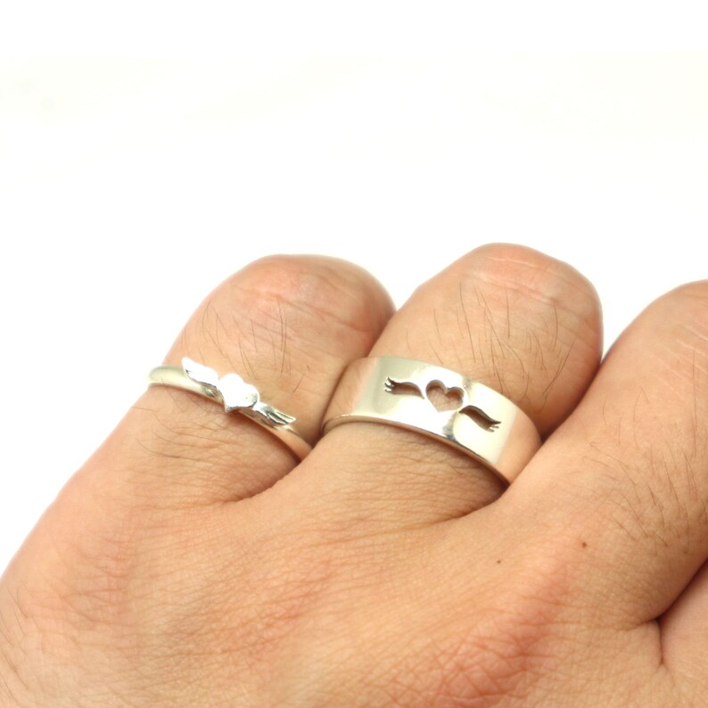 May include: Two silver rings with a heart design. The ring on the left has a small heart with wings, while the ring on the right has a larger heart with wings on either side.