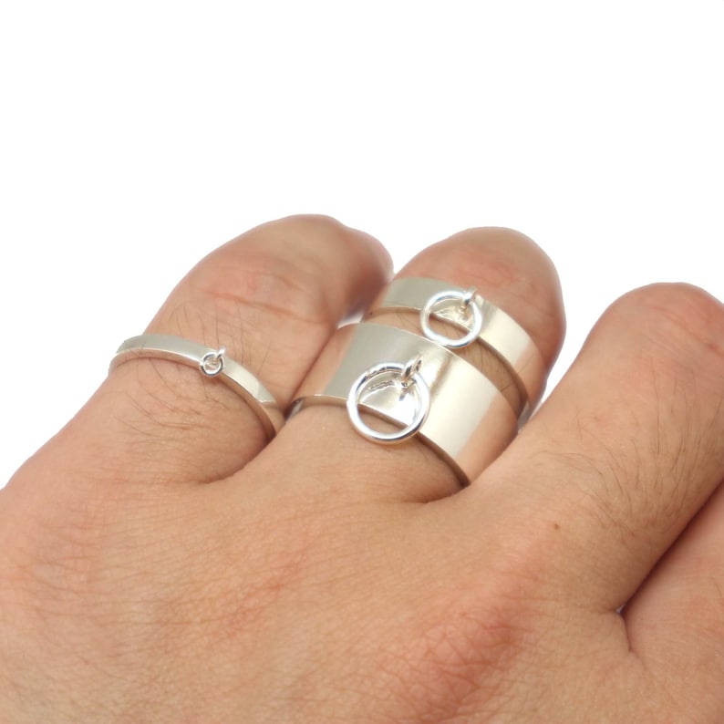 May include: Three silver rings with a simple design. The two larger rings have a small circle attached to the band. The rings are worn on a person's finger.