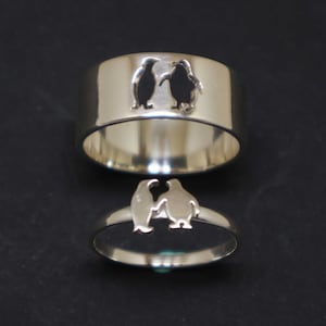Könnte beinhalten: Zwei silberne Ringe mit einem Ausschnitt-Design von zwei sich gegenüber stehenden Pinguinen. Die Ringe sind übereinander gestapelt.
