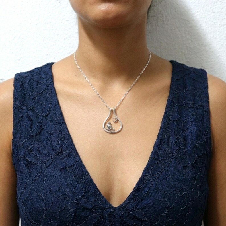 May include: A silver pendant necklace with a teardrop design, featuring a small bird and a gemstone. The necklace is worn by a person wearing a navy blue dress.