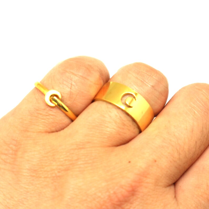 May include: Two gold rings on a hand. The larger ring has a cut-out letter "C" and the smaller ring is a simple band with a crescent moon shape.