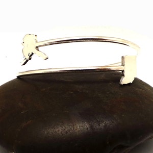 May include: Silver bangle bracelet with a cutout of the state of Alaska on one side and the state of Indiana on the other side. The bracelet is resting on a dark brown rock.