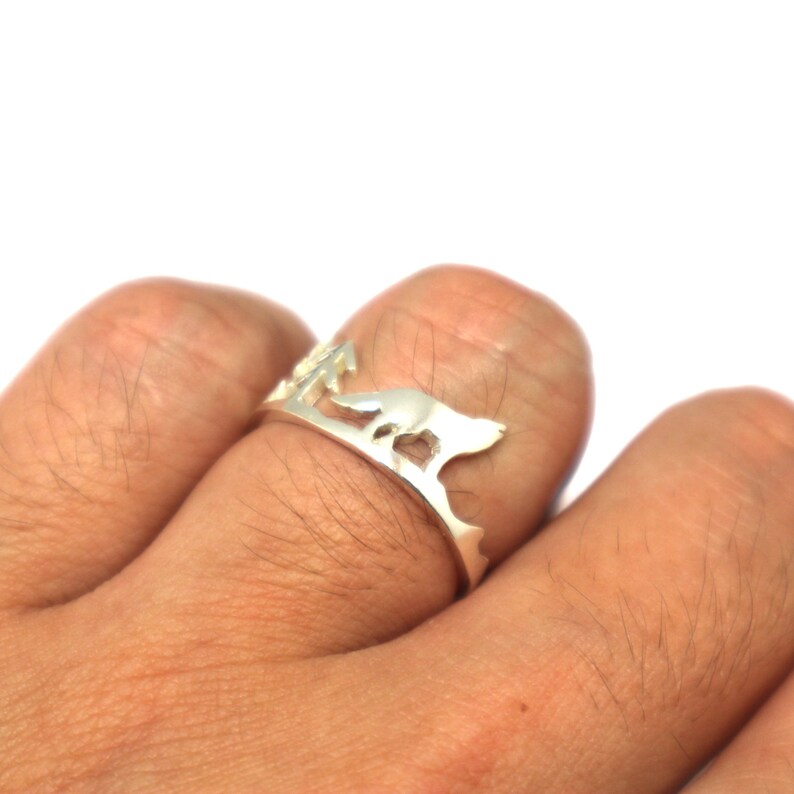 May include: A silver ring featuring a cut-out design of a wolf running through a forest. The ring is worn on a person's finger.