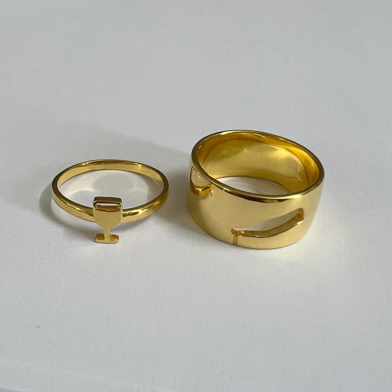 May include: Two gold rings, one with a simple design and the other with a cutout shape. The rings are on a white surface.