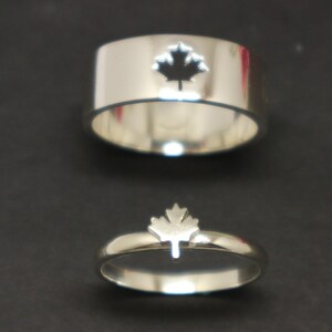 Puede incluir: Dos anillos de plata con diseño de hoja de arce. Uno es una banda ancha con un recorte de hoja de arce. El otro es una banda más delgada con forma de hoja de arce. Ambos anillos están sobre un fondo oscuro.