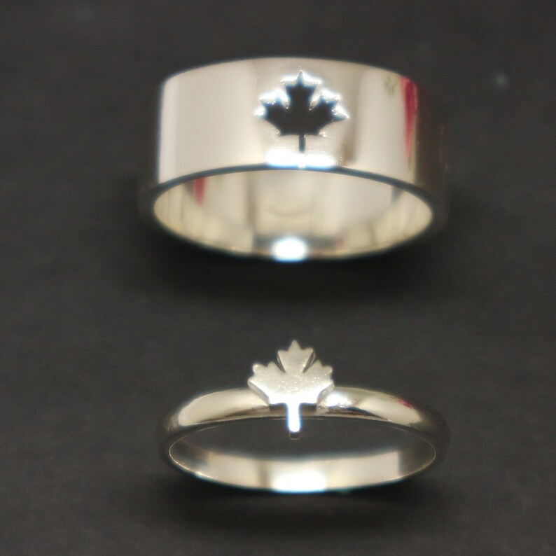 May include: Two silver rings with a maple leaf design. One ring is a wide band with a maple leaf cutout. The other ring is a thinner band with a maple leaf shape on top. Both rings are on a dark background.