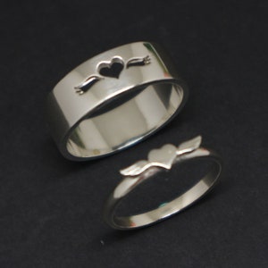 May include: Two silver rings with a heart design. One ring has a heart with wings and the other has a heart with a banner.