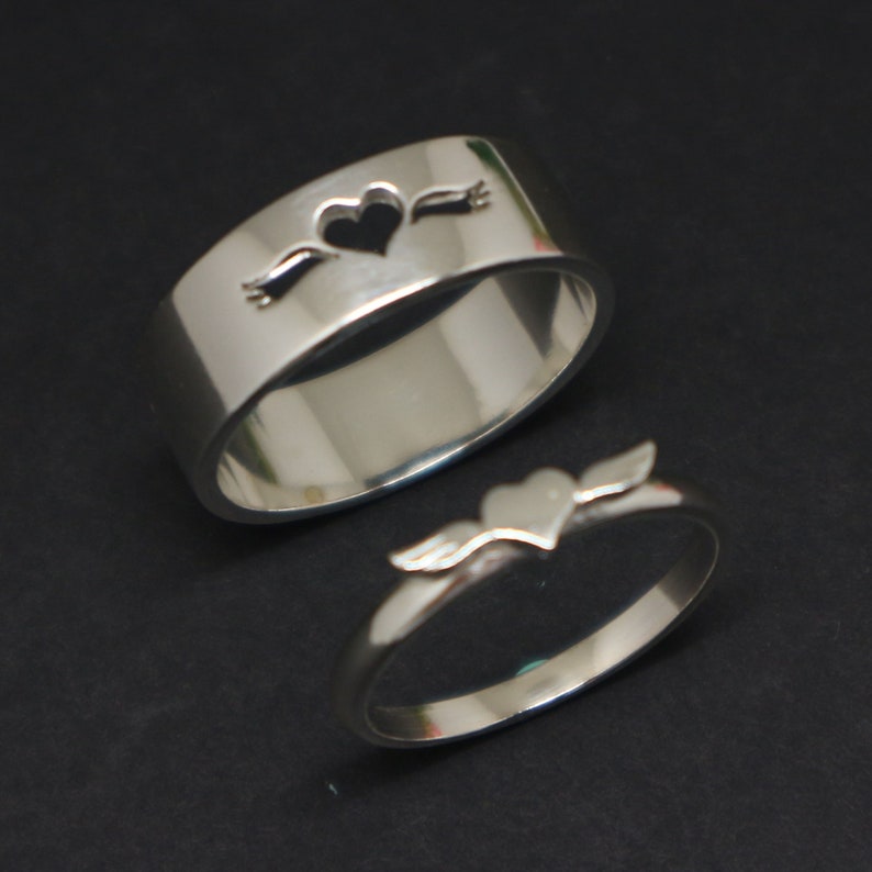 May include: Two silver rings with a heart design. One ring has a heart with wings and the other has a heart with a banner.