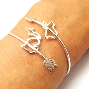 Peut inclure: Bracelet en argent avec une découpe de l'état du Michigan et du Texas, avec une flèche pointant vers la droite. Le bracelet est porté au poignet d'une personne.