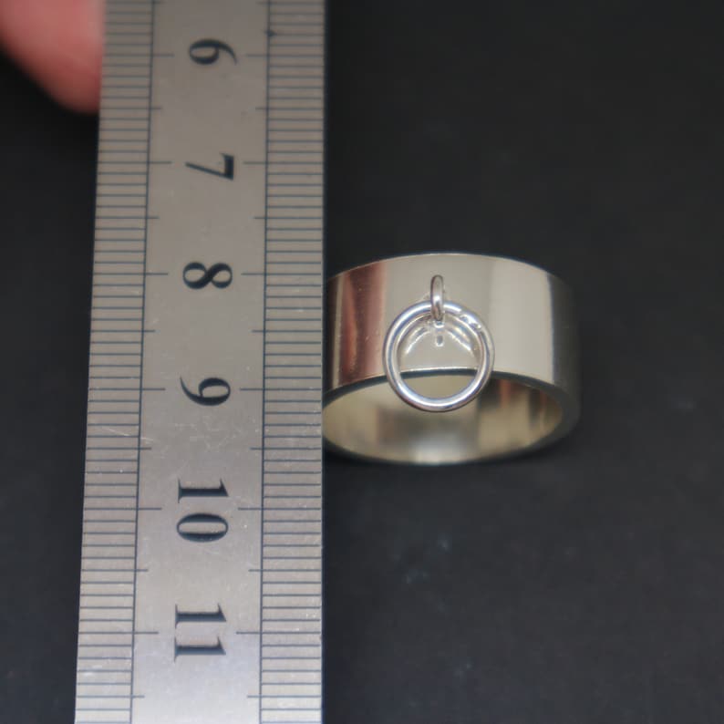 May include: A silver ring with a thick band and a small silver ring attached to the top. The ring is measured against a ruler.