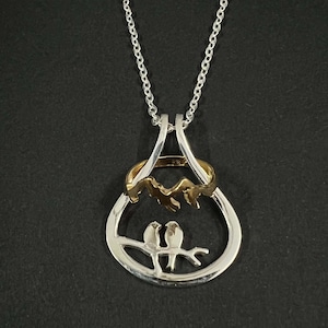 May include: Silver necklace with a pendant featuring two birds perched on a branch. The pendant is made of two interlocking rings, one silver and one gold, with a mountain range design.