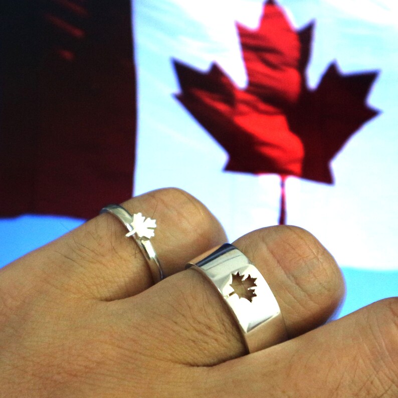 May include: Two silver rings, one with a small maple leaf design and the other with a cut-out maple leaf. The rings are displayed against a backdrop of the Canadian flag, featuring a red maple leaf on a white background.