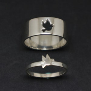 Op de afbeelding: Twee zilveren ringen met een duivenontwerp. Een ring heeft een uitgesneden duif en de andere ring heeft een massieve duif.