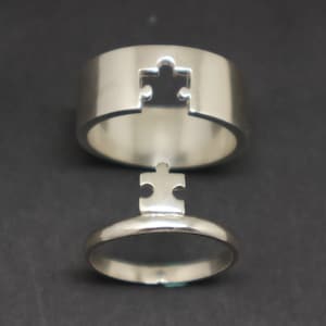 May include: Two silver puzzle piece rings. The top ring has a puzzle piece cut out of it. The bottom ring has a puzzle piece that fits into the top ring.