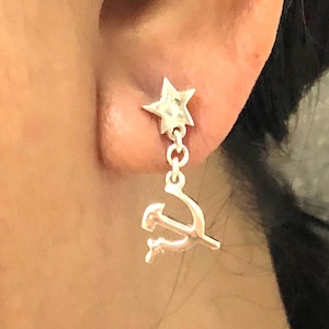 Puede incluir: Pendiente plateado con una estrella como perno y un dije colgante de martillo y hoz. El pendiente está diseñado con un eslabón de cadena. El pendiente se lleva en la oreja.