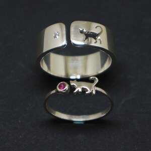 May include: Two silver rings with cat silhouettes. The top ring has a single diamond and the bottom ring has a pink gemstone.